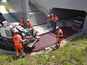 Robotnicy obsługują maszynę drogową kładącą barwiony na czerwono asfalt u wlotu ścieżki rowerowej do przejścia podziemnego.