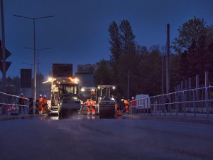 Nocne asfaltowanie ul. Cieszyńskiej. Ujęcie z niskiej perspektywy. Na tle dymiącej od ciepła masy bitumicznej zespół drogowy wykonuje prace asfaltowe.