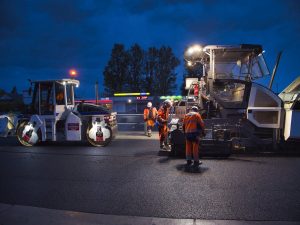 Zdjęcie nocne. Widok na ekipę drogowców obsługujących park maszynowy do robót bitumicznych w trakcie asfaltowania.