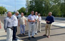 Siedem osób stoi przy drodze i rozmawia; w tle chodnik, latarnie, znaki i drzewa w słoneczny dzień.