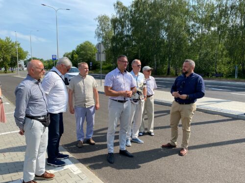 Siedem osób stoi przy drodze i rozmawia; w tle chodnik, latarnie, znaki i drzewa w słoneczny dzień.
