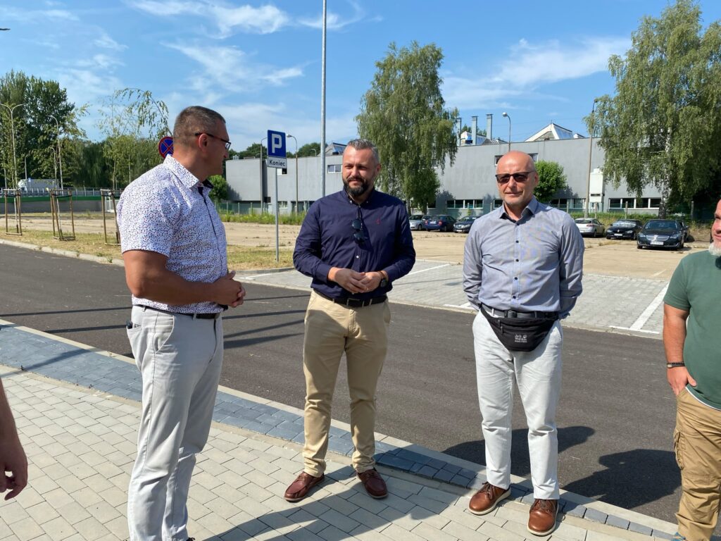 Cztery osoby stoją obok parkingu; w tle zaparkowane auta, budynek i znak „Parking - Koniec”.