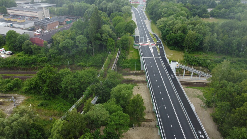 Widok z góry na drogowy most nad torami kolejowymi, z oznaczeniami pasów i ścieżki rowerowej, otoczony drzewami.